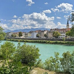 Draupromenade 12 Villach – turquoise river Drau with the old town roofline, church spire and mountains behind
