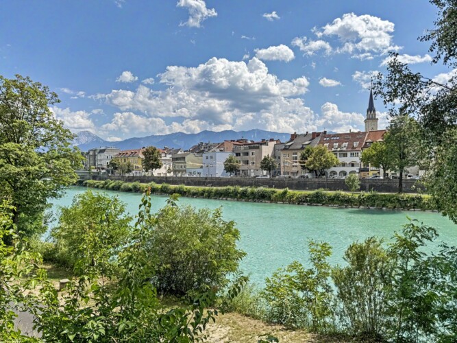 Draupromenade 12 Villach – turquoise river Drau with the old town roofline, church spire and mountains behind