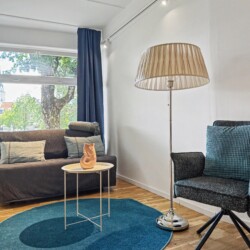 Draupromenade 12 Villach – Top 1 Türkis – sofa bed, tall floor lamp and dark swivel chair on the round turquoise rug