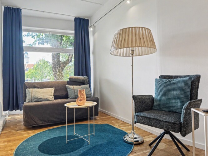 Draupromenade 12 Villach – Top 1 Türkis – sofa bed, tall floor lamp and dark swivel chair on the round turquoise rug