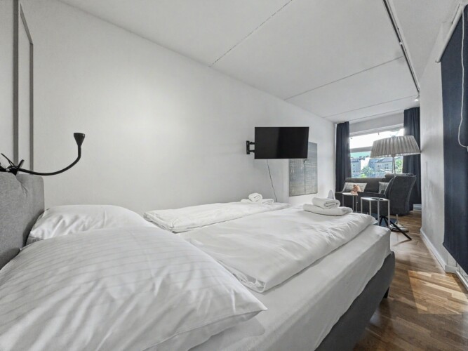 Draupromenade 12 Villach – Top 1 Türkis – king bed with upholstered headboard, black reading lamp and wall-mounted TV