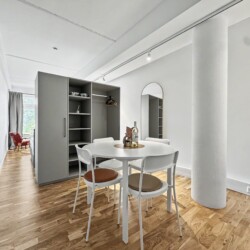 Draupromenade 12 Villach – Top 2 Gold – open-plan room with round dining table, exposed concrete column and open wardrobe
