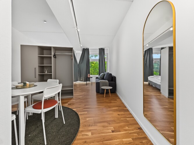 Draupromenade 12 Villach – Top 3 Drau – open-plan room with round dining table, built-in wardrobe, sofa at the window and gold arched full-length mirror