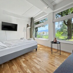 Draupromenade 12 Villach – Top 3 Drau – king bed lengthways along the panorama window with wall-mounted TV and sofa