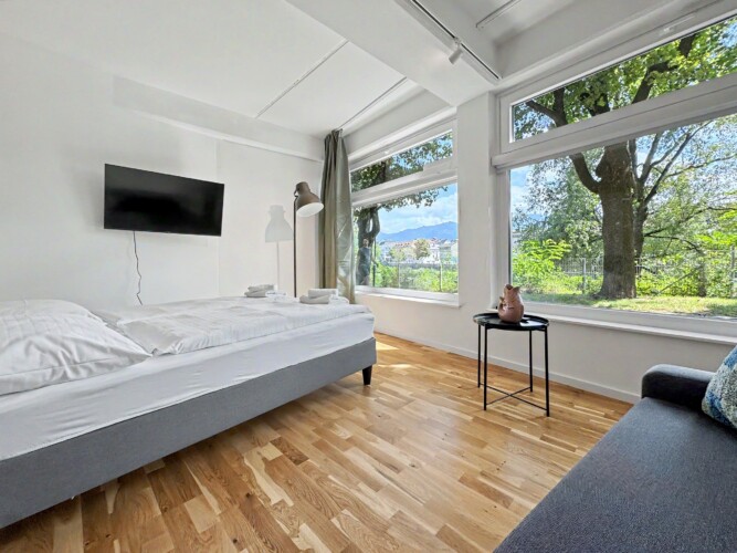 Draupromenade 12 Villach – Top 3 Drau – king bed lengthways along the panorama window with wall-mounted TV and sofa