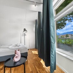 Draupromenade 12 Villach – Top 3 Drau – petrol-green blackout curtains at the panorama window next to the bed with black floor lamp