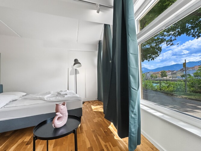 Draupromenade 12 Villach – Top 3 Drau – petrol-green blackout curtains at the panorama window next to the bed with black floor lamp