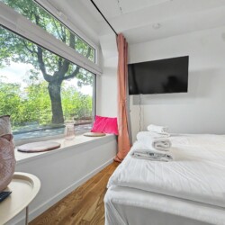 Draupromenade 12 Villach – Top 4 Pink – king bed facing the wall-mounted TV, window nook with pink cushion and glass vase
