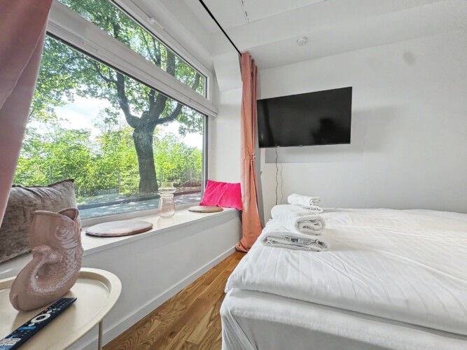 Draupromenade 12 Villach – Top 4 Pink – king bed facing the wall-mounted TV, window nook with pink cushion and glass vase